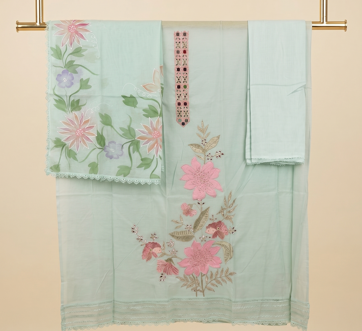 A mint green embroidered traditional outfit set hanging on a gold rod against a beige background. The set includes a kurta with pink and green floral embroidery, a matching dupatta with pink flowers, and a folded matching fabric piece.