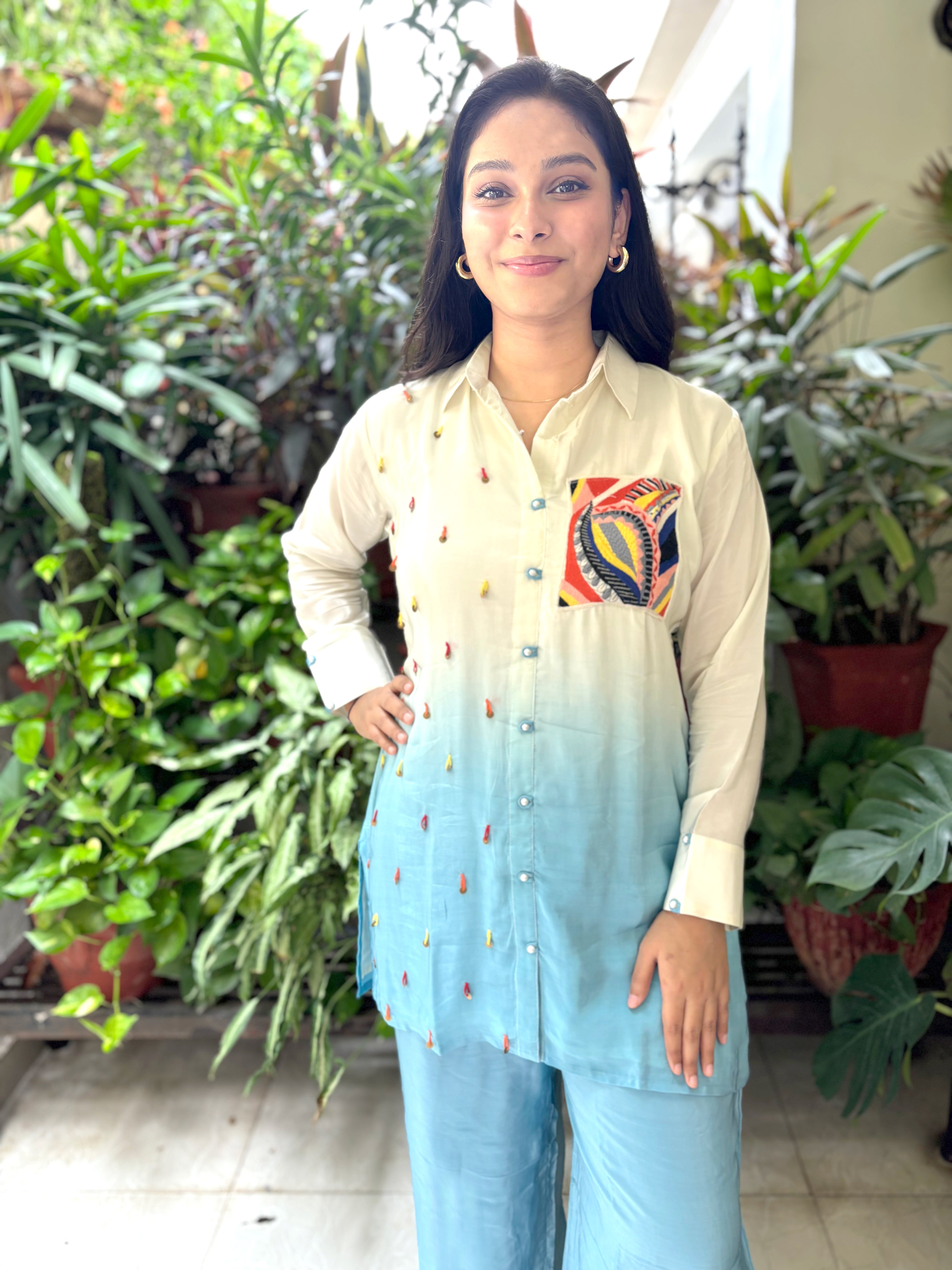 A woman modeling a light beige to sky blue ombre kurta with colorful embroidered details and a patterned pocket, paired with matching light blue pants, set against a background of potted green plants.