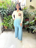 A woman stands in a tiled indoor space surrounded by potted plants, wearing a cream and light blue gradient button-up shirt with colorful embroidered details on the chest, paired with wide-leg light blue jeans.