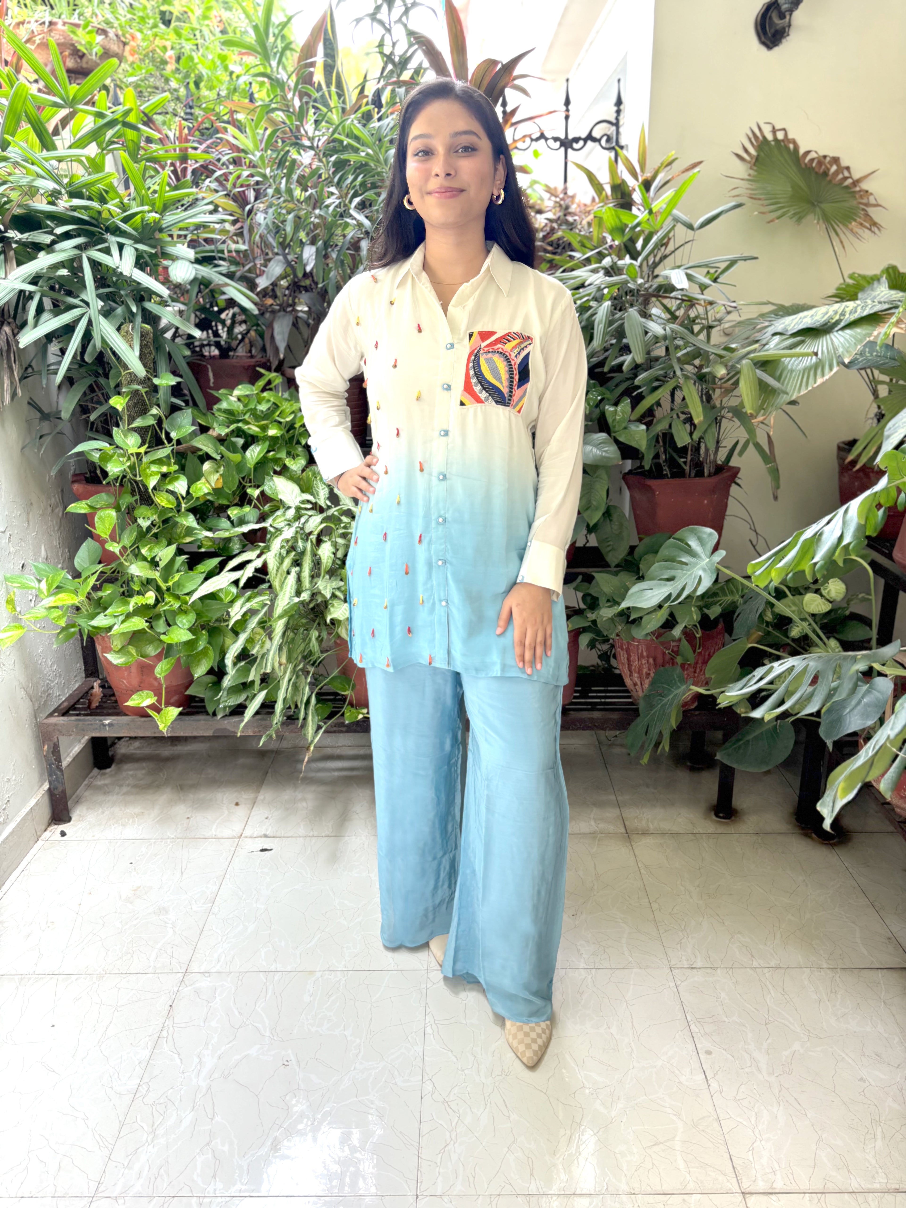 A woman stands in a tiled indoor space surrounded by potted plants, wearing a cream and light blue gradient button-up shirt with colorful embroidered details on the chest, paired with wide-leg light blue jeans.