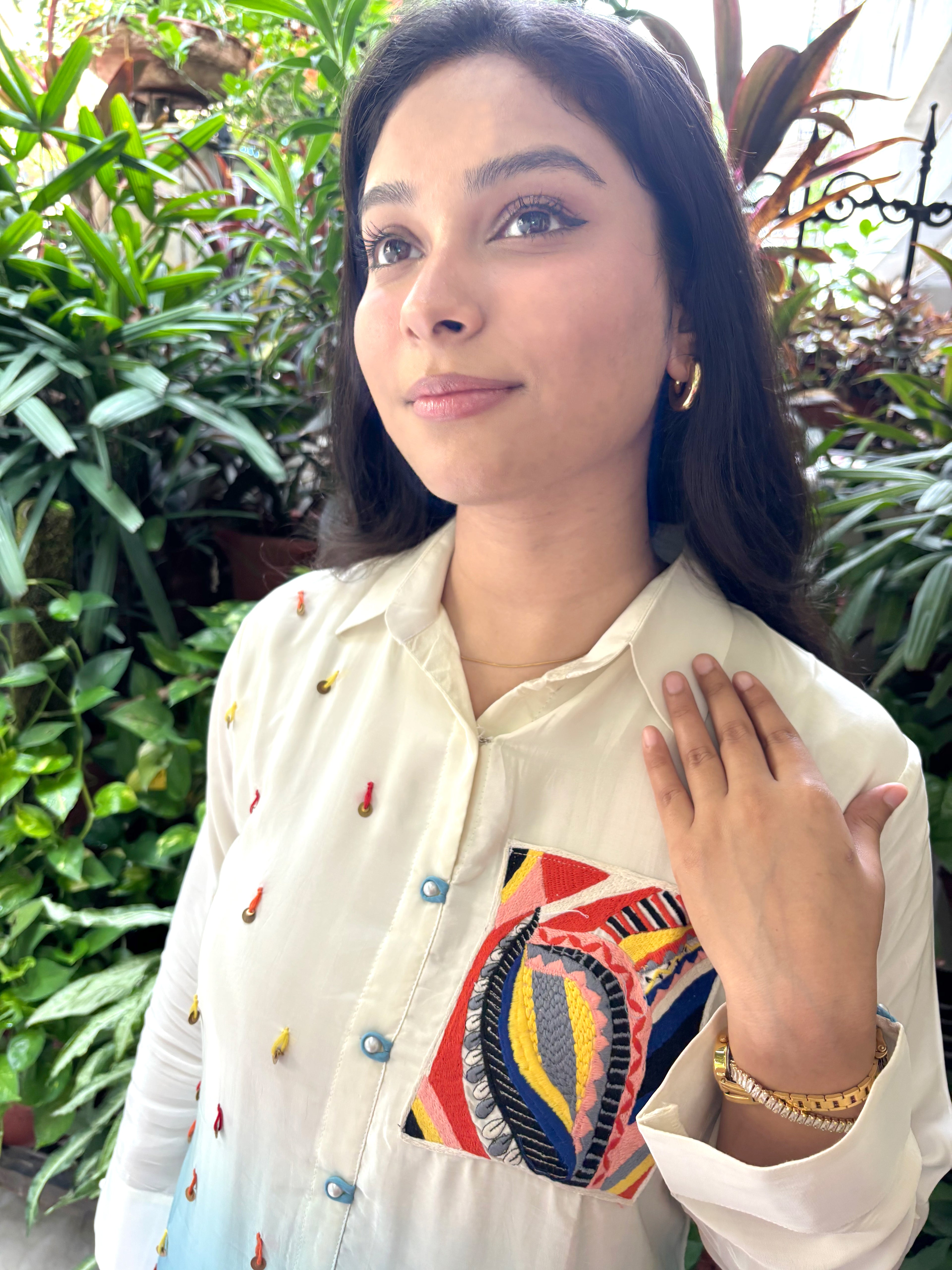 A woman wearing a cream-colored button-up shirt with colorful geometric embroidery on the chest pocket and small embroidered details on the sleeves, posing in front of a lush green plant background.