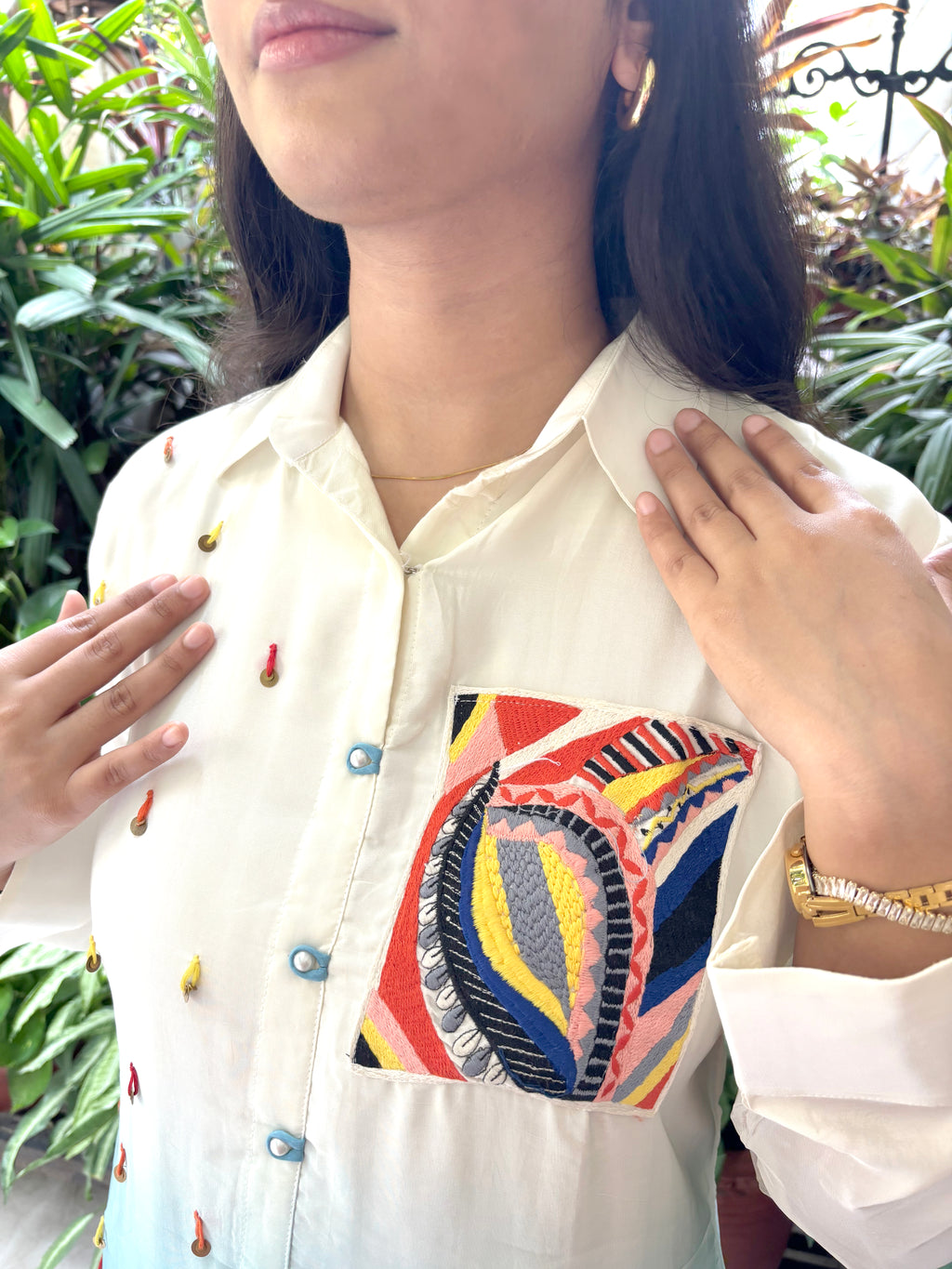 A close-up of a woman wearing a cream-colored button-up shirt with colorful embroidered details on the chest pocket and small embroidered accents on the sleeves. The background shows green foliage.