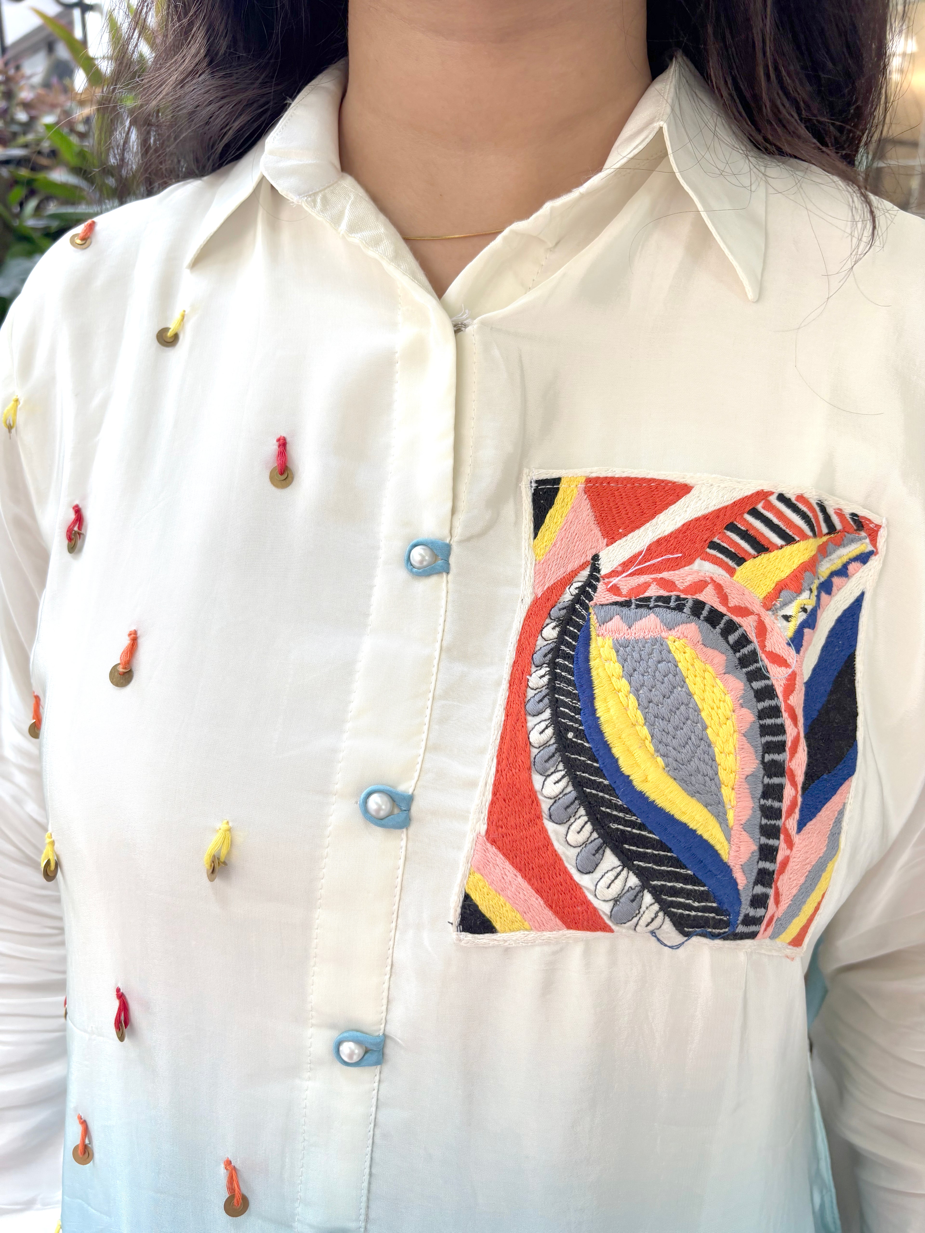 A close-up of a person wearing a cream-colored long-sleeve button-up shirt with colorful embroidered details on the chest pocket and scattered small embroidered accents on the front. The pocket features a large, vibrant paisley-style embroidery in red, yellow, blue, black, and white.