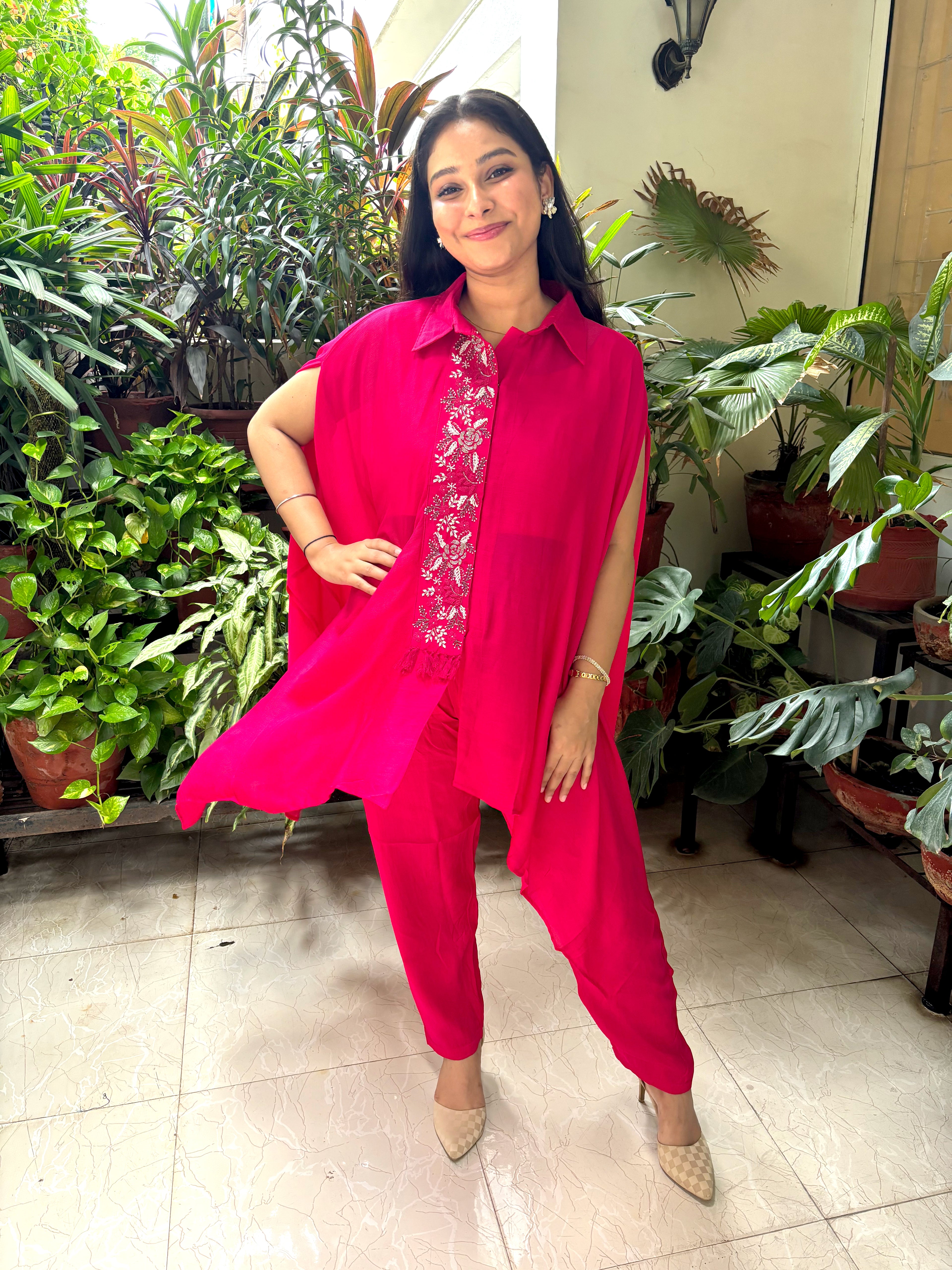 A woman modeling a bright fuchsia pink three-piece traditional outfit consisting of a sleeveless button-up shirt, matching pants, and a white embroidered inner blouse, standing in a tiled indoor space surrounded by potted green plants.
