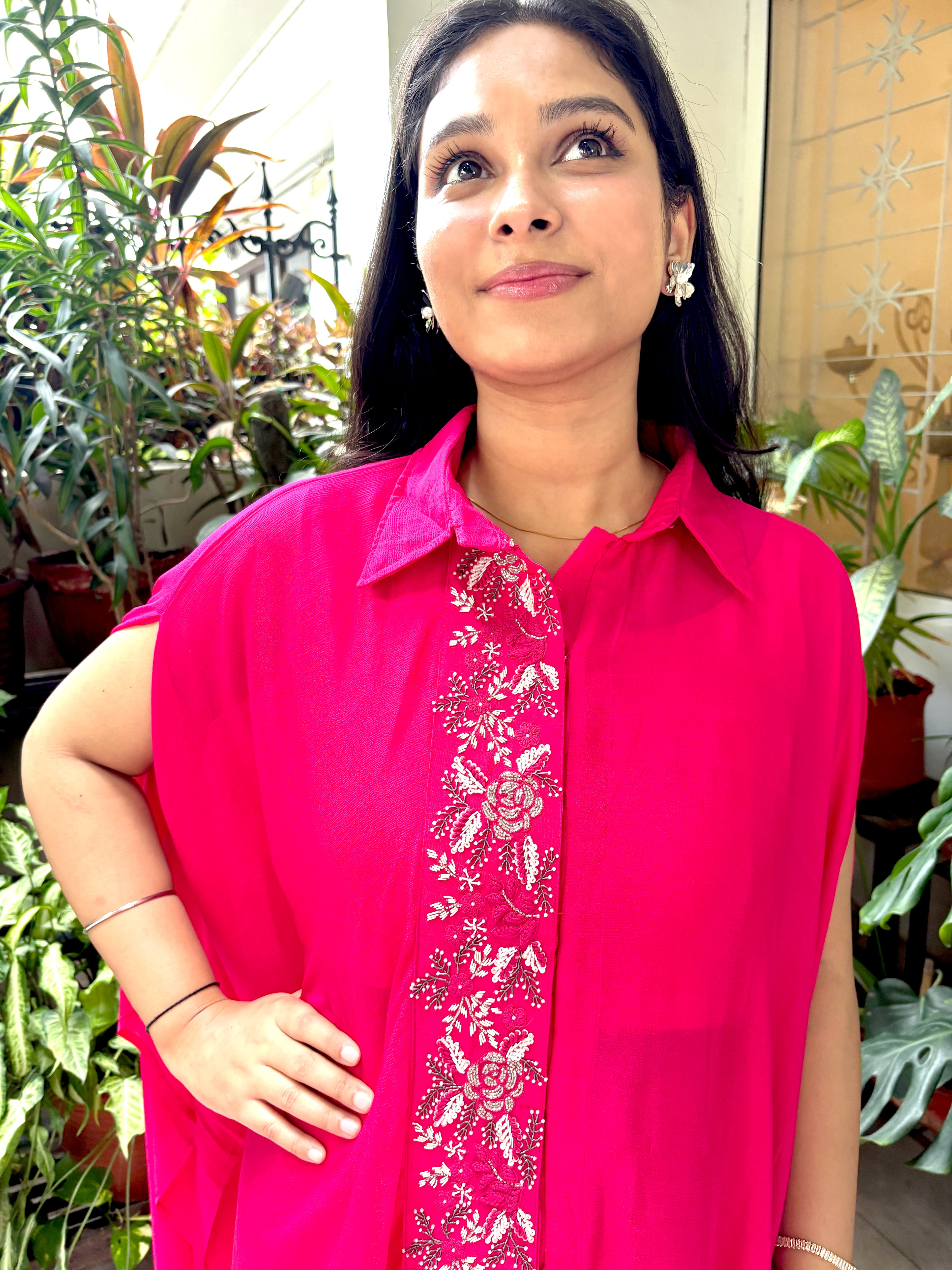 A woman wearing a bright pink sleeveless button-up shirt with a matching embroidered front panel, posed in front of a background of indoor plants.