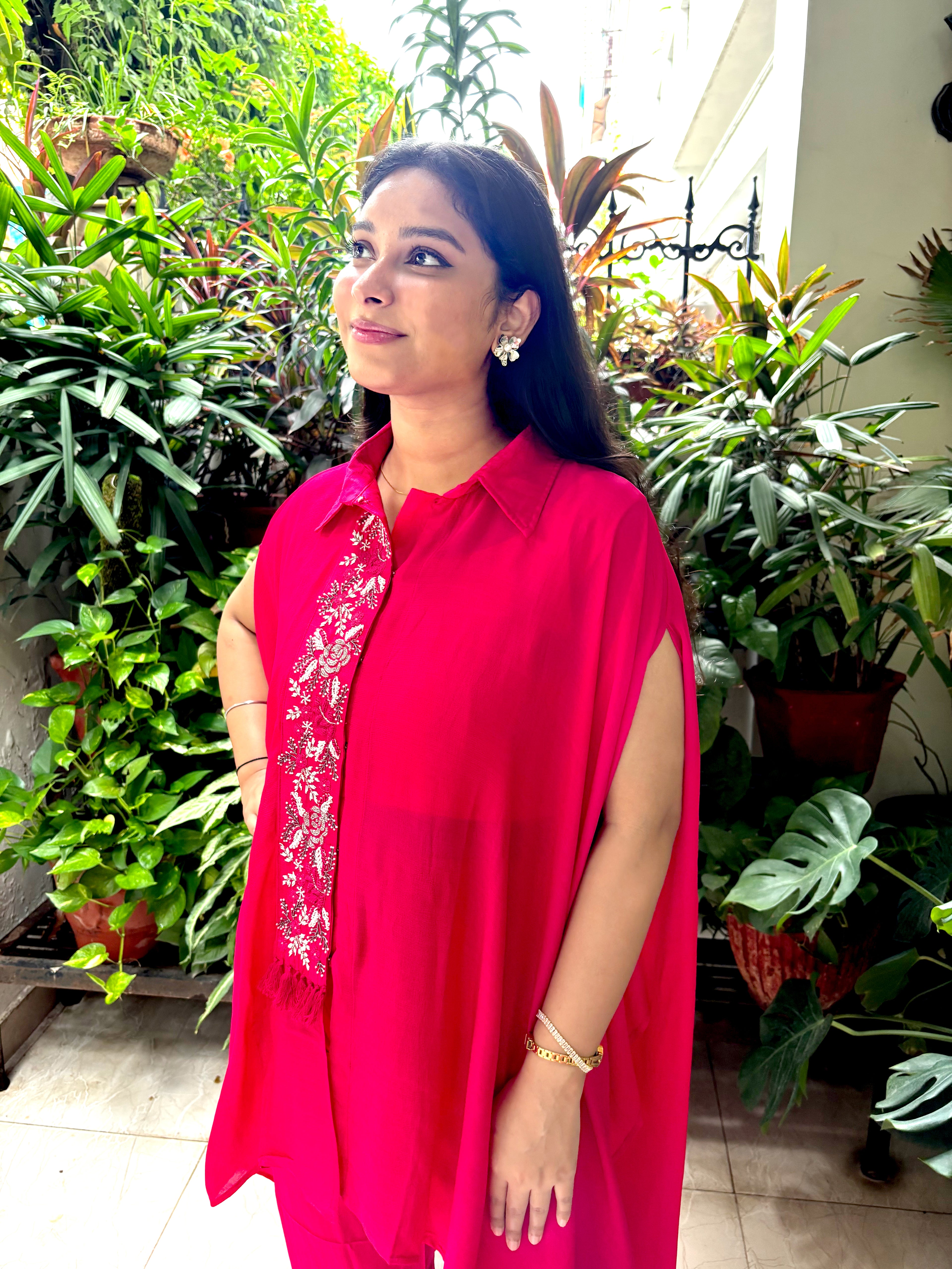 A woman wearing a bright pink sleeveless tunic top with a matching pink scarf featuring white floral embroidery, standing in a lush indoor garden setting.