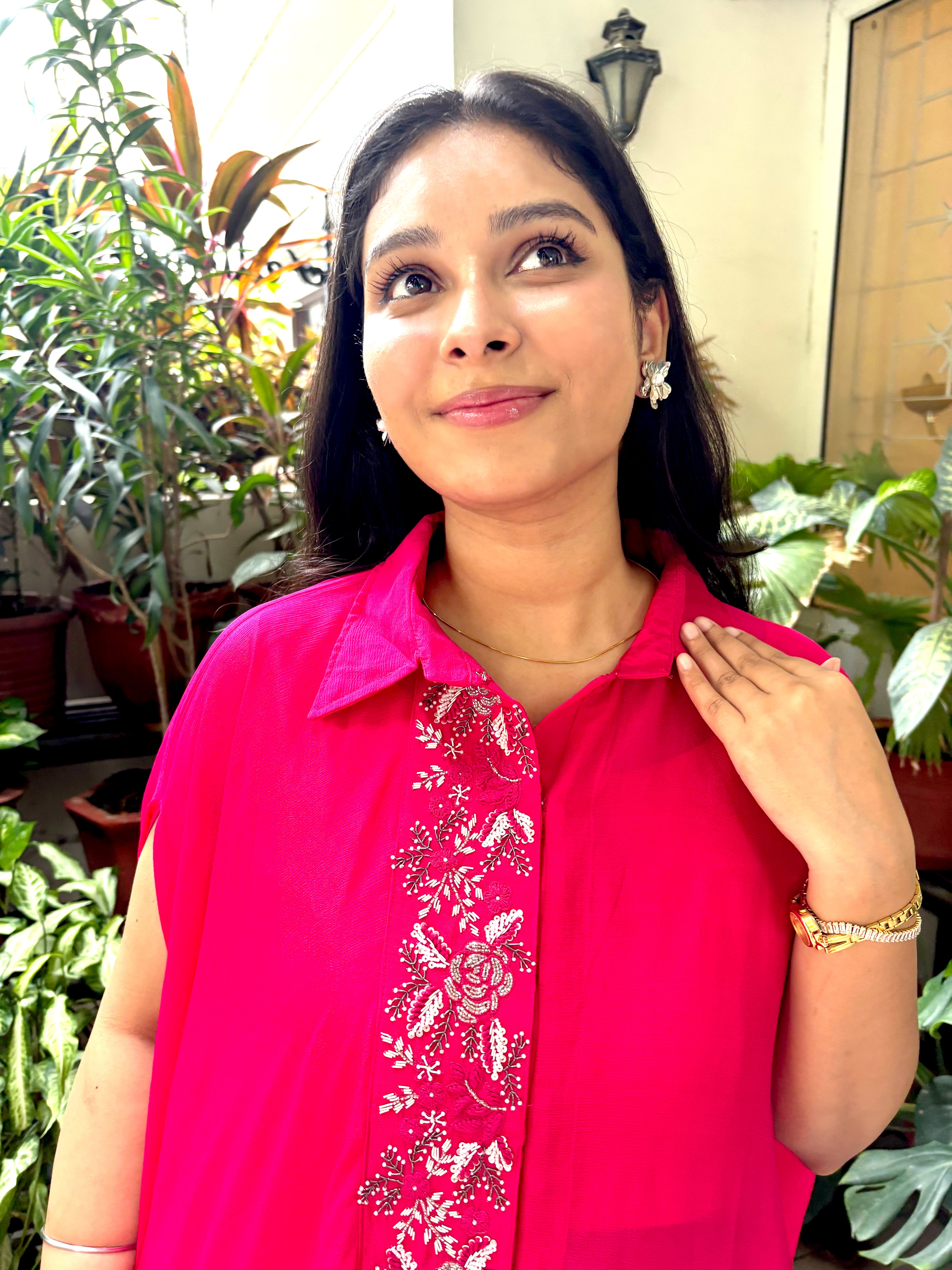 A woman wearing a bright pink sleeveless collared top with white floral embroidery down the front, standing in front of a background of potted green plants.