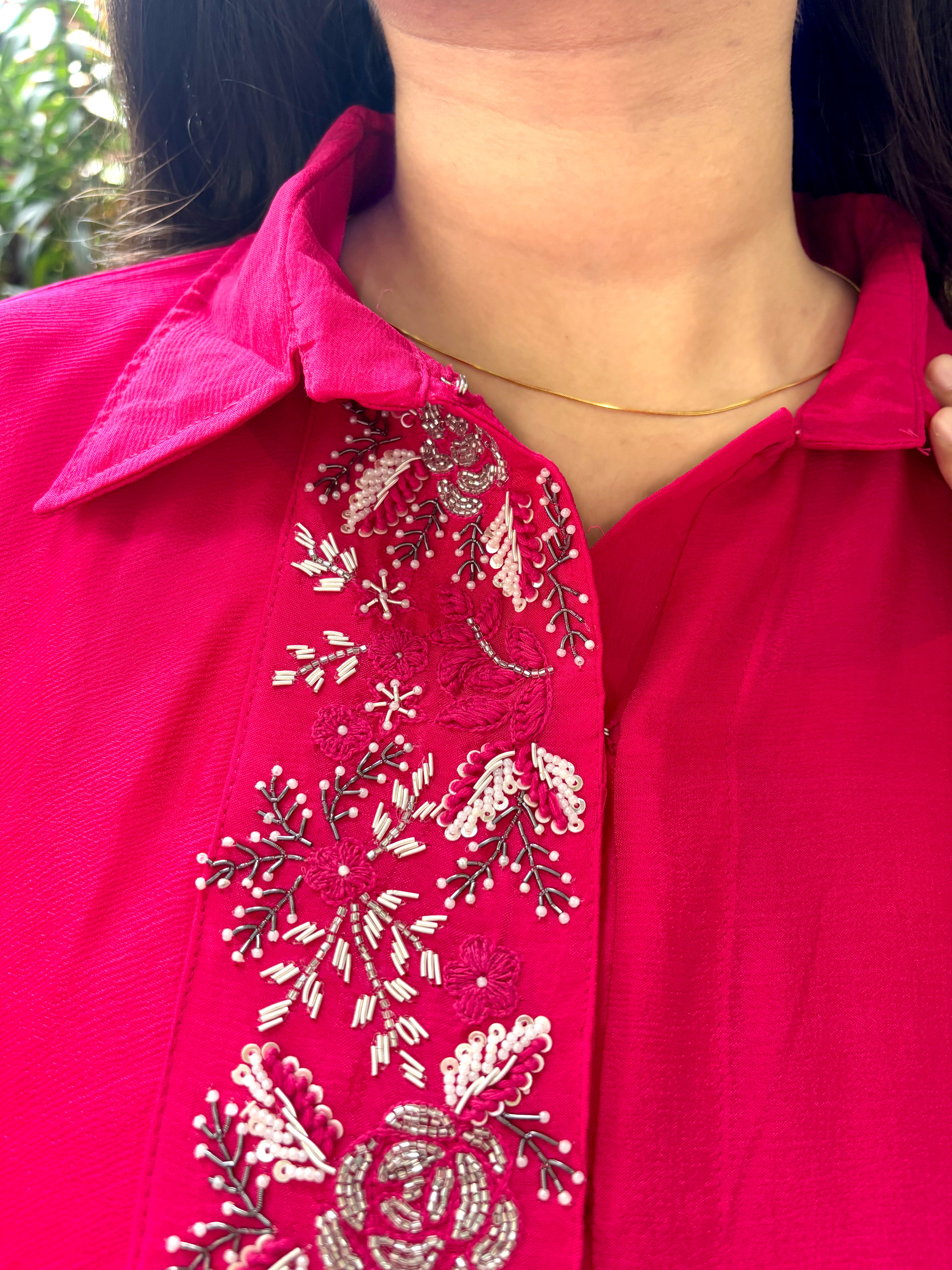 A close-up of a person wearing a bright pink collared blouse with ornate silver and white floral embroidery down the front placket.
