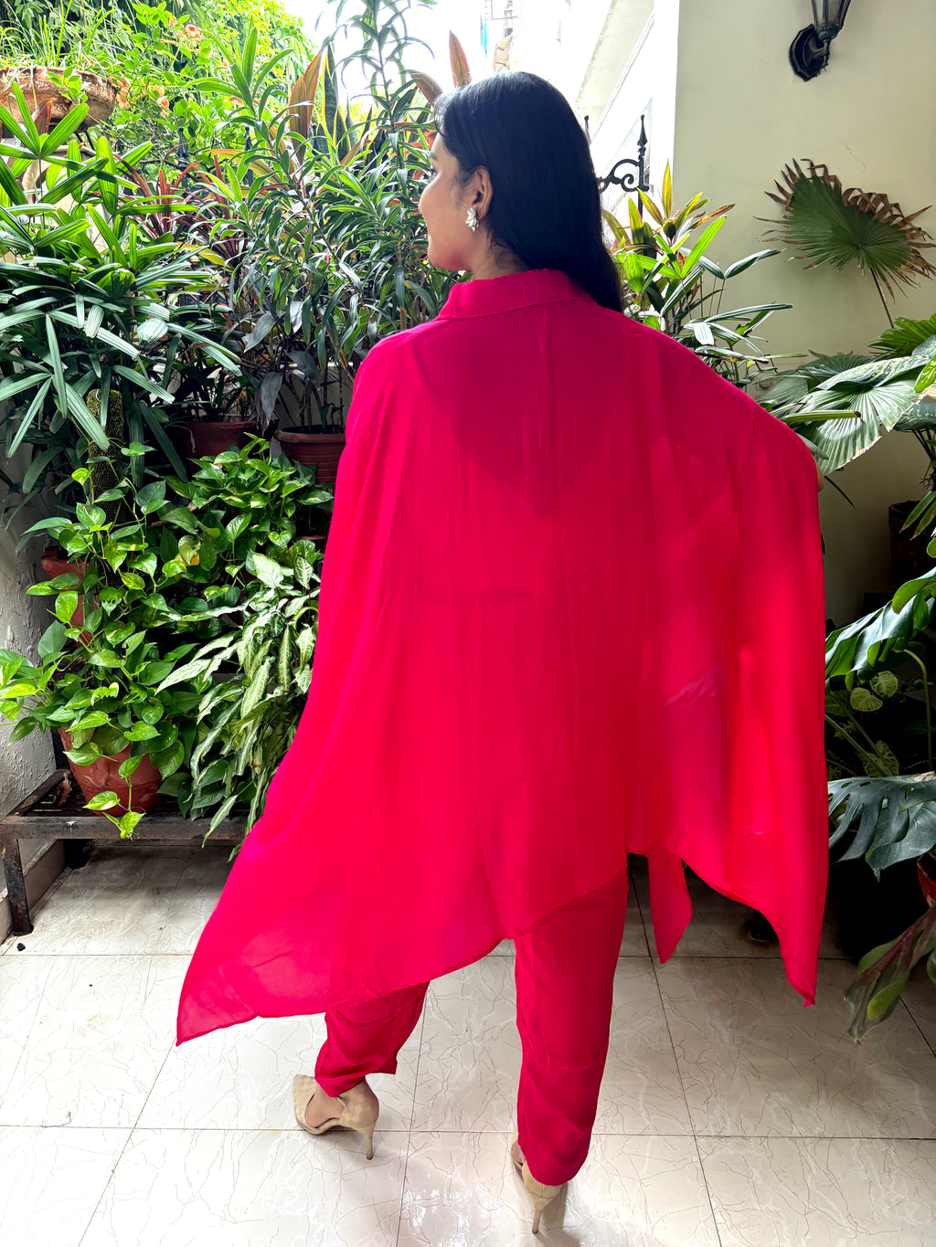 A woman wearing a bright red matching outfit, featuring a loose draped cape-style top and matching pants, photographed from behind in an indoor garden setting with potted plants.