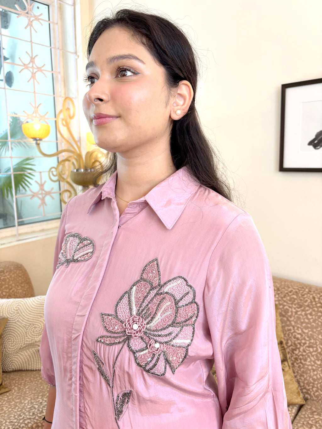 A woman wearing a light pink long-sleeve button-up shirt with a large floral embroidery design on the front, standing in a living room setting.