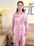 A woman wearing a light pink three-piece kurta set with floral embroidery, standing in a living room with patterned sofas and a lamp in the background.