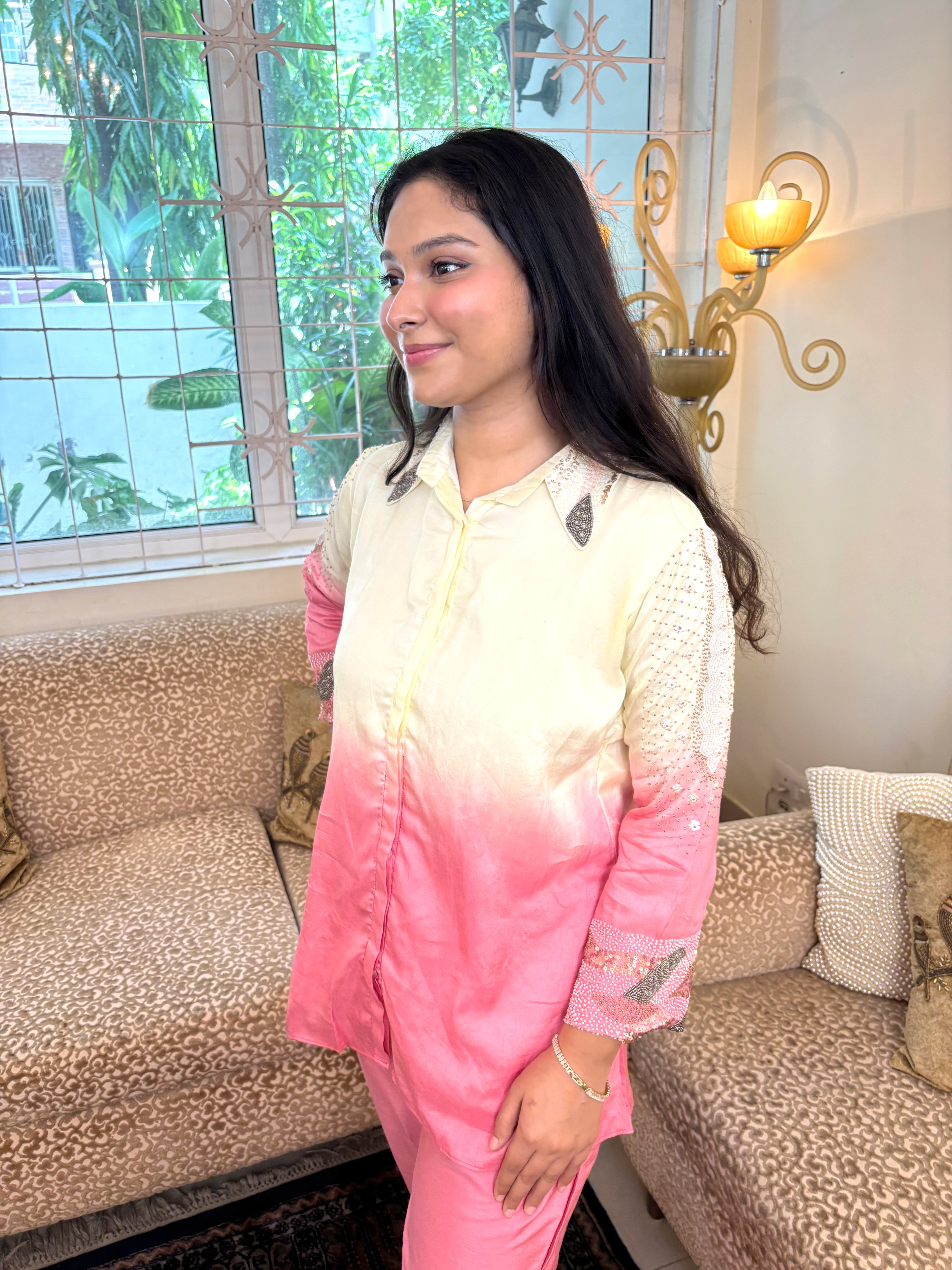 A woman wearing a pink and cream ombré pajama set with lace sleeve details, standing in a living room in front of a window and sofa.