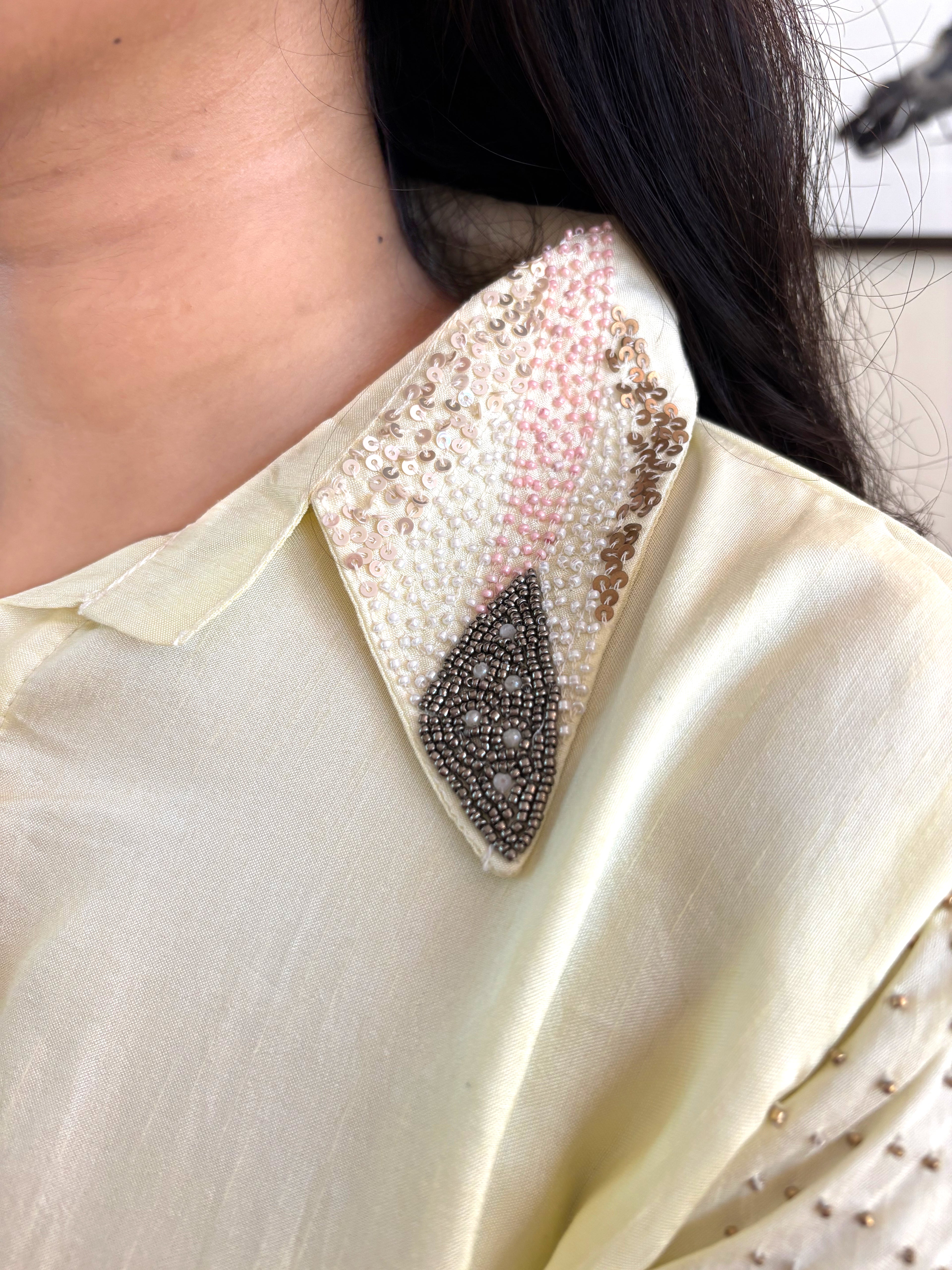 Close-up of a person wearing a cream-colored blouse with a decorative pointed collar embellished with white pearls, pink sequins, and a dark gray beaded geometric motif.