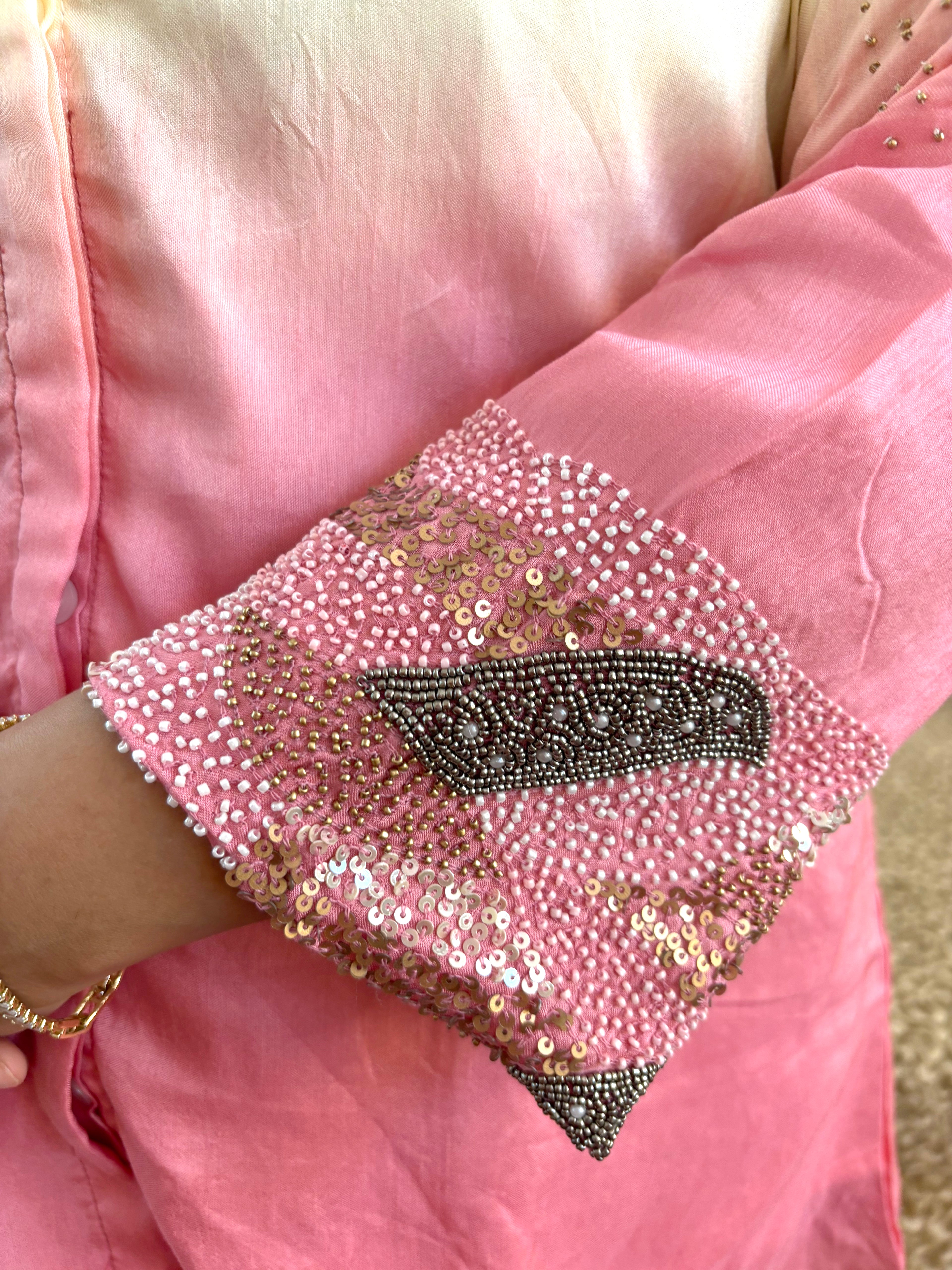 A close-up of a pink traditional garment sleeve decorated with dense pink, gold, and dark gray sequins and beadwork, worn on a person's arm.