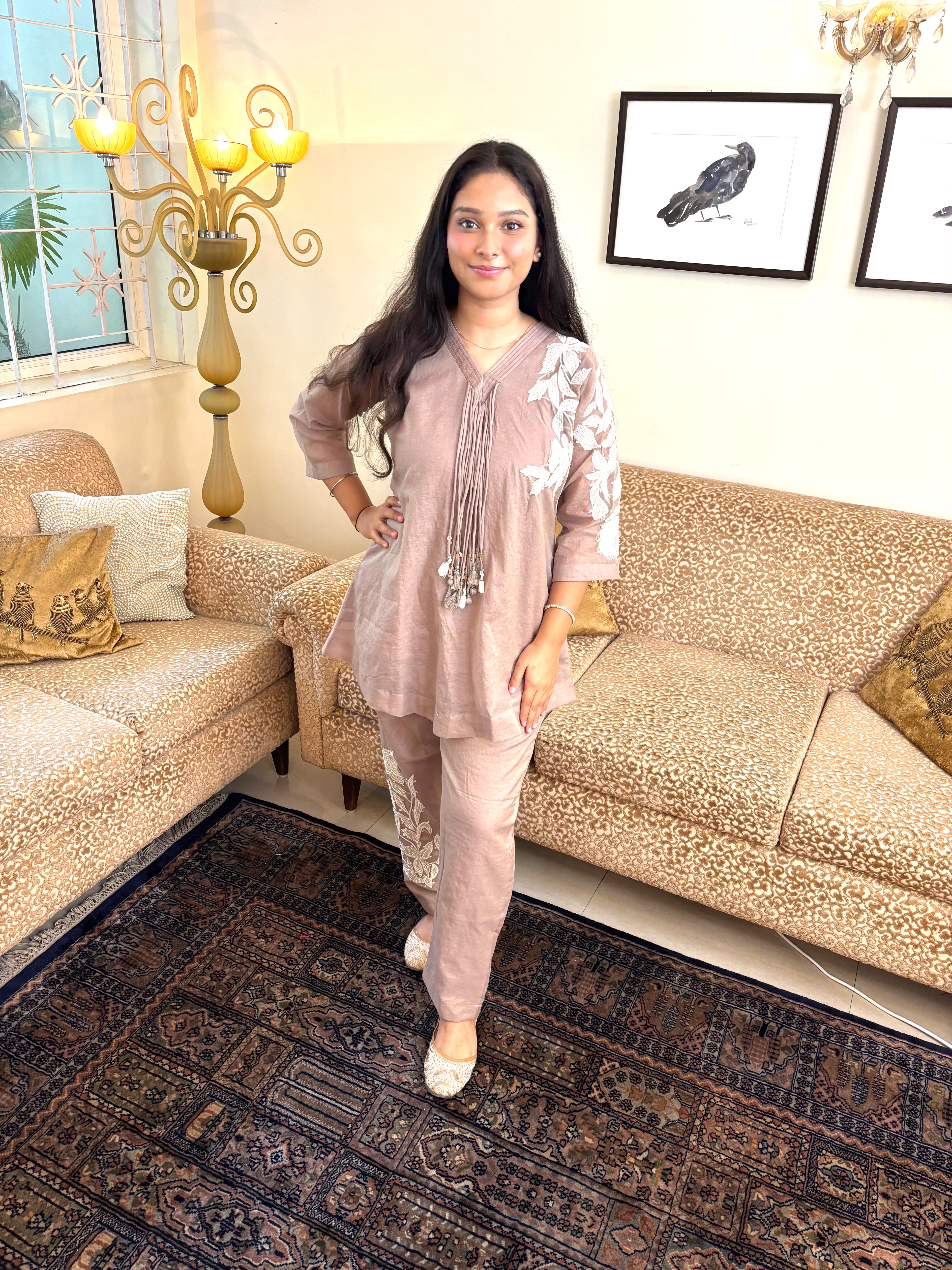 A woman modeling a light pink two-piece traditional outfit with white floral embroidery on the sleeves, standing in a decorated living room with patterned sofas and a dark rug.
