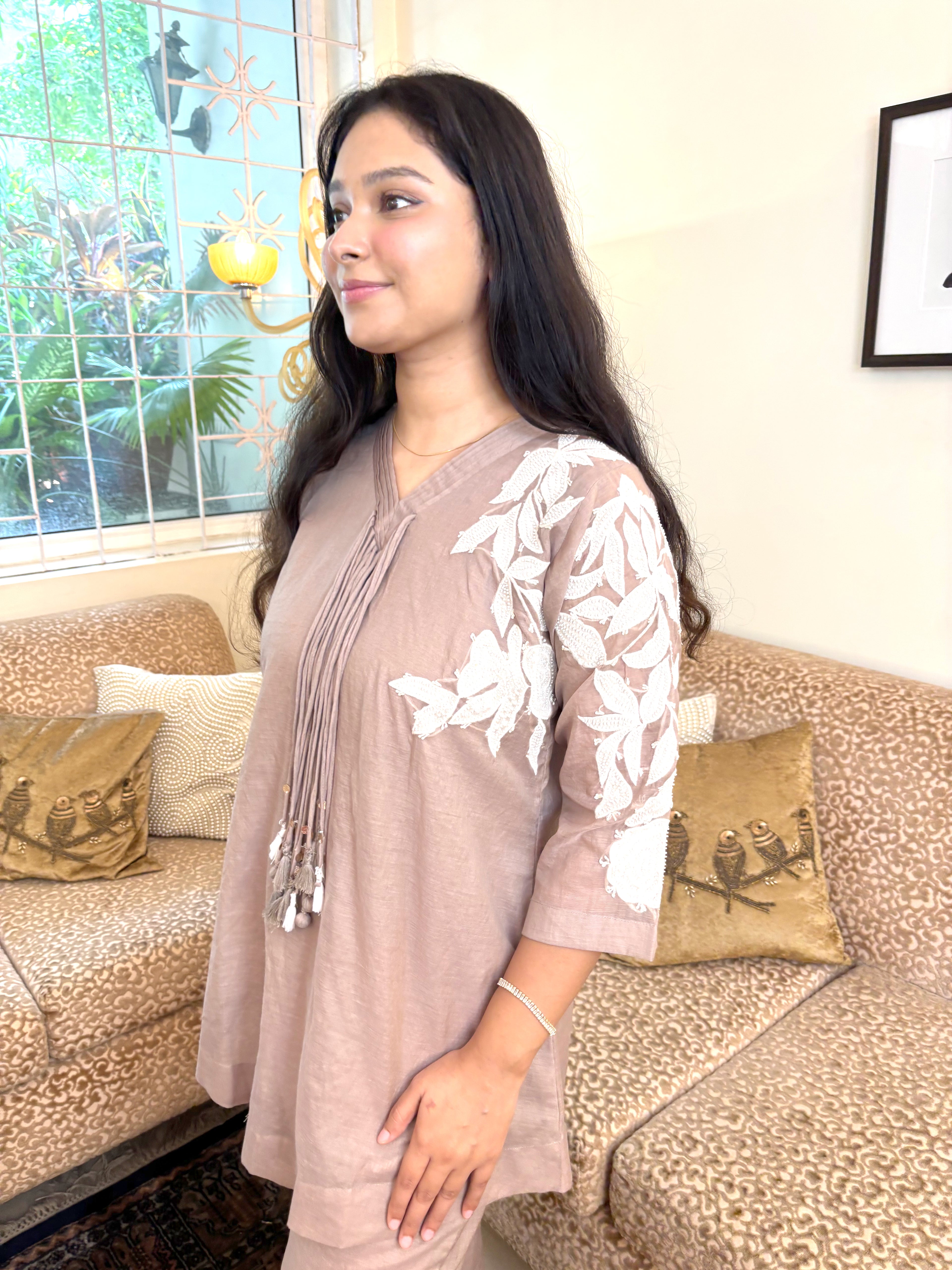A woman wearing a taupe-beige tunic top with white floral embroidery on the sleeves, standing in a living room in front of a patterned sofa.