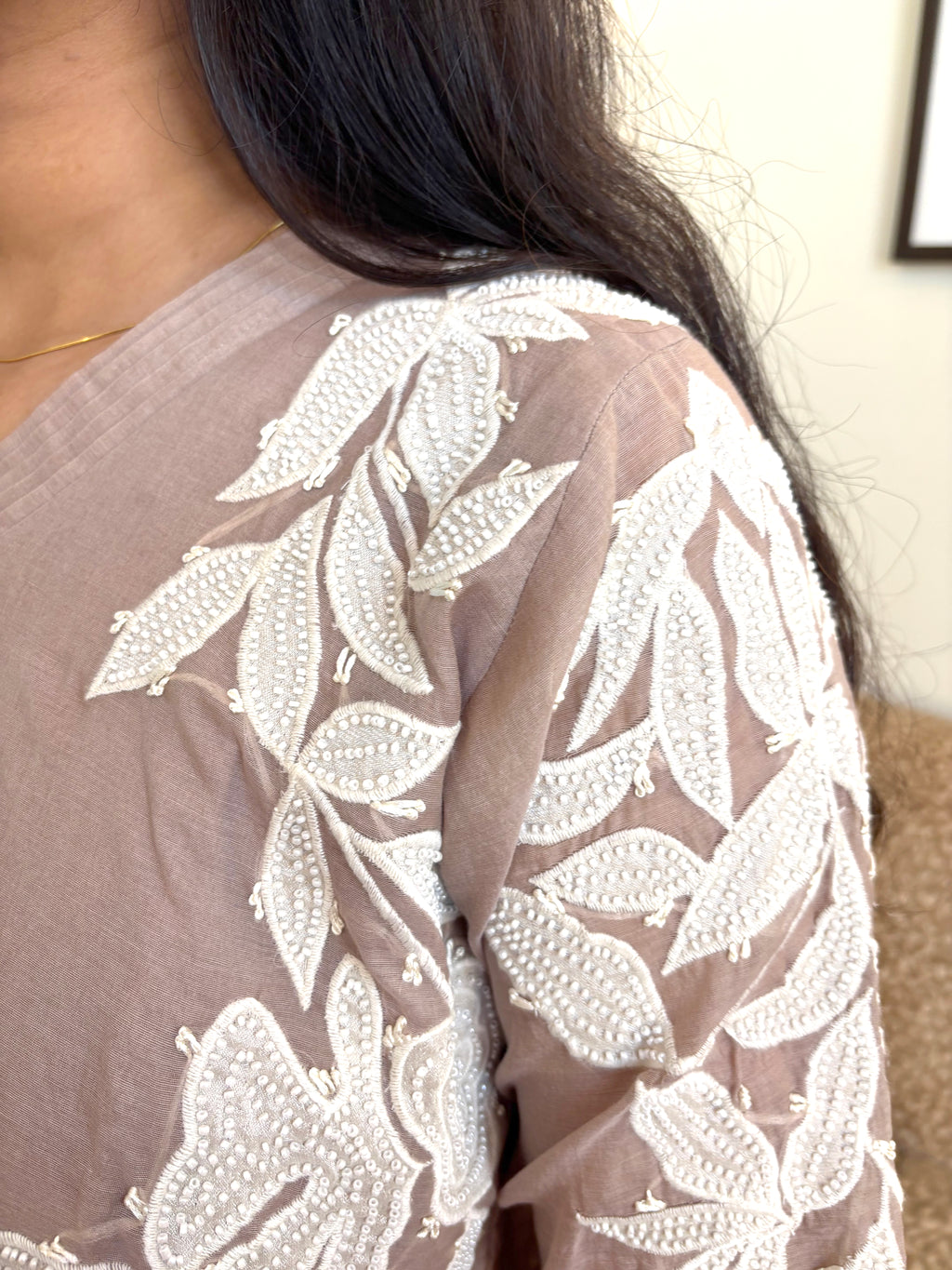 Close-up of a person wearing a sheer taupe-brown top with intricate white floral embroidery and beadwork on the shoulder and sleeve.
