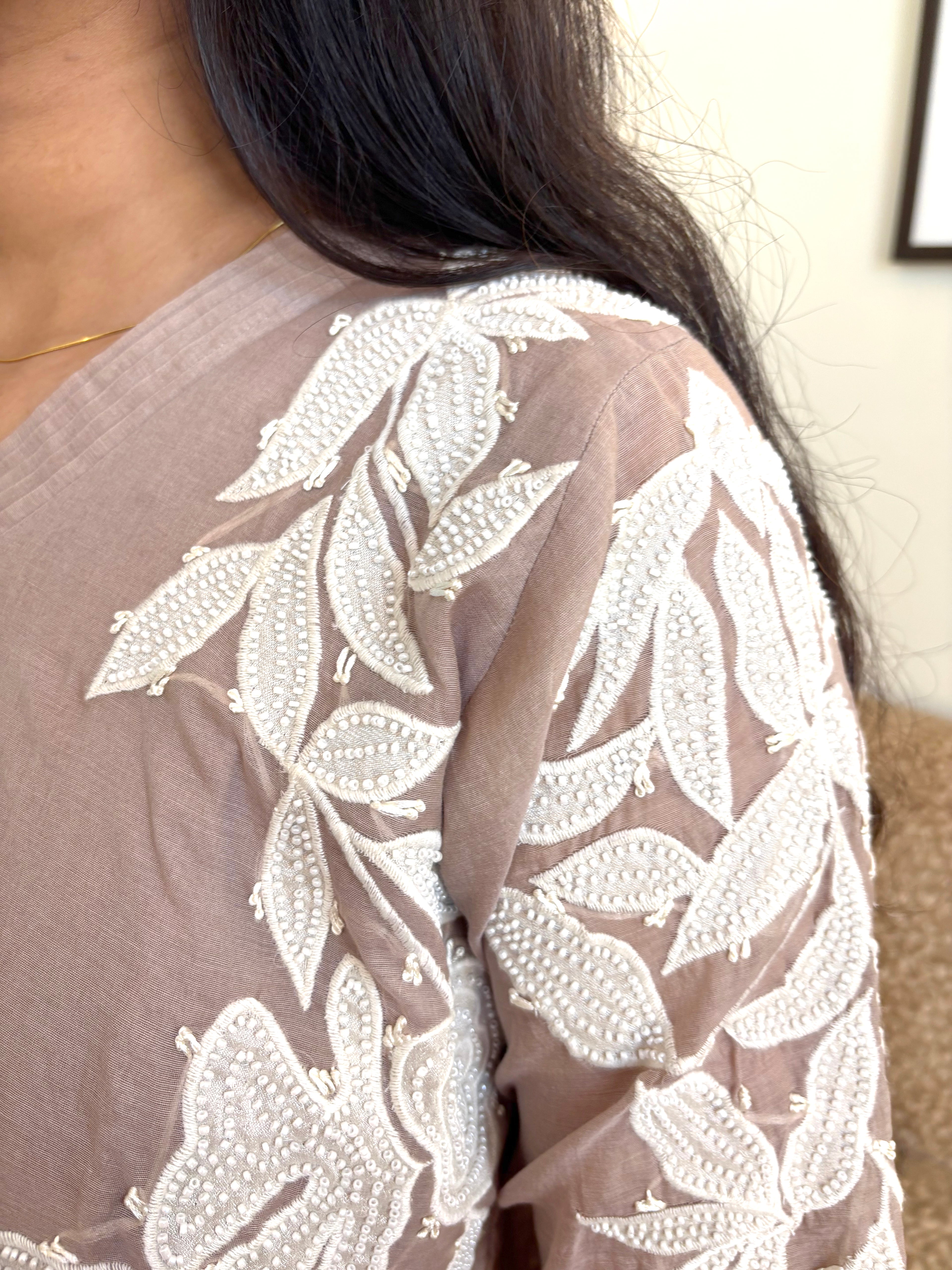 Close-up of a person wearing a sheer taupe-brown top with intricate white floral embroidery and beadwork on the shoulder and sleeve.