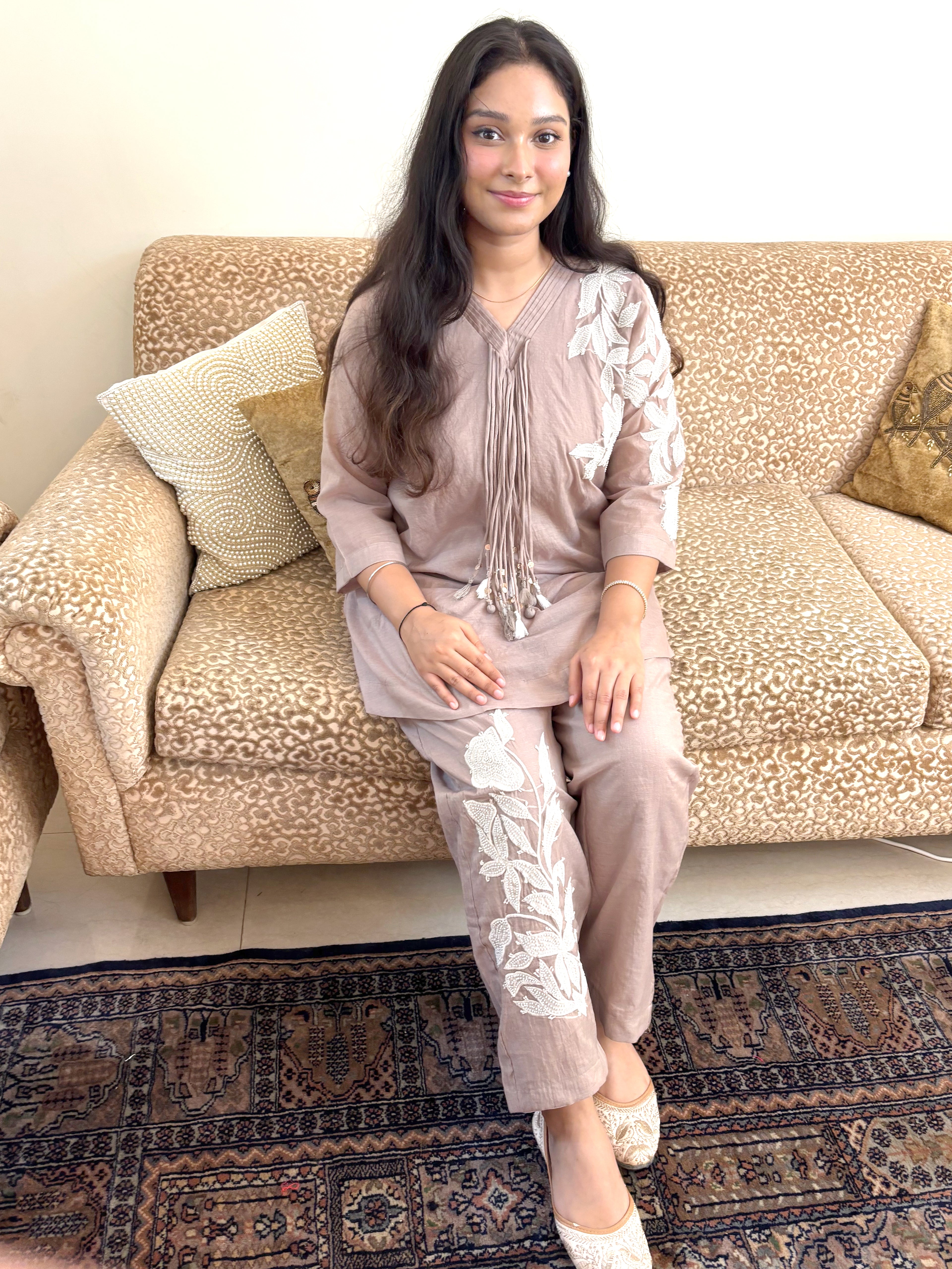 A woman wearing a matching beige and white printed kurta set sits on a patterned sofa in a living room setting.