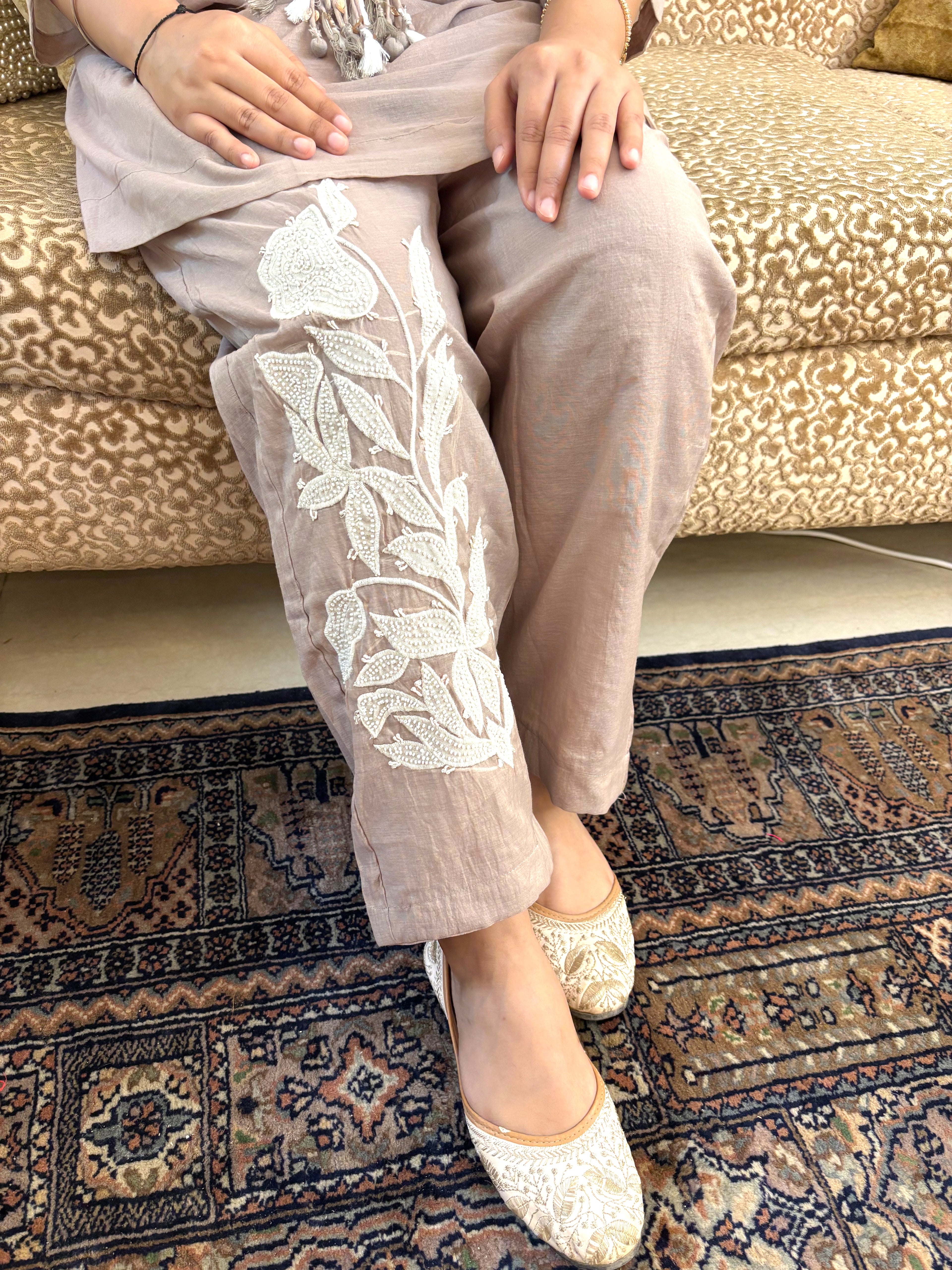 A person seated on a patterned sofa wearing taupe-beige pants with a large white floral embroidery down one leg, paired with white lace flat shoes and a patterned rug underneath.