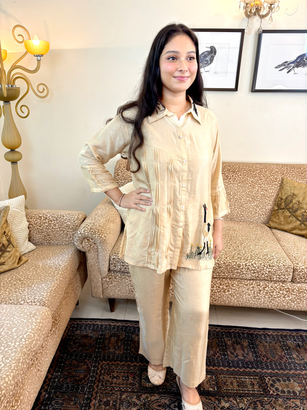 A woman modeling a light beige two-piece pajama set consisting of a long-sleeve button-up top and matching pants, standing in a living room with patterned sofas and a rug.