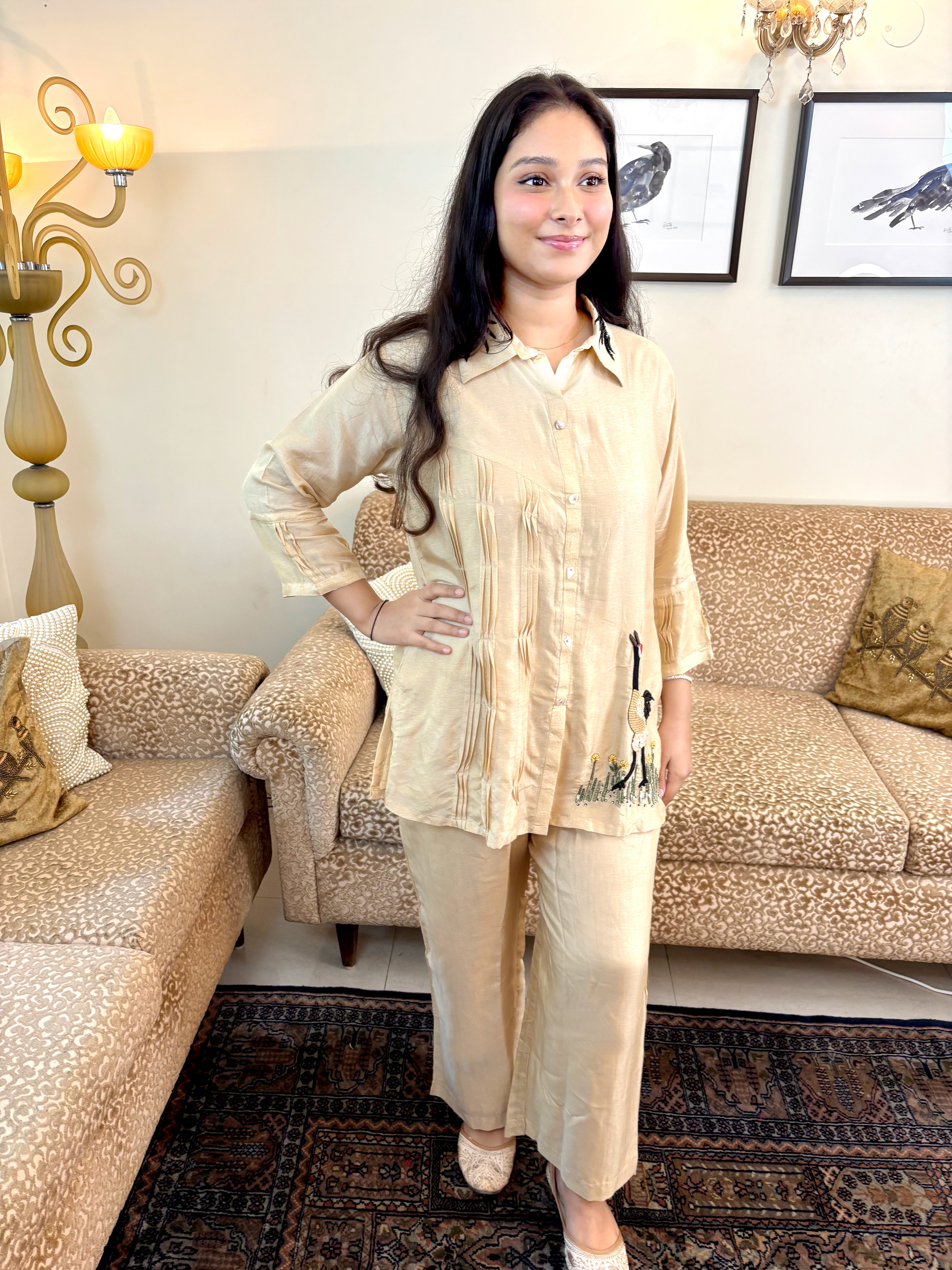 A woman modeling a light beige two-piece pajama set consisting of a long-sleeve button-up top and matching pants, standing in a living room with patterned sofas and a rug.