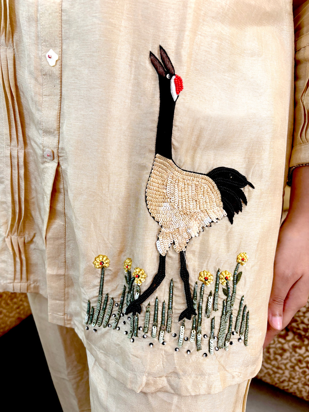 A close-up of a beige embroidered tunic or kurta featuring a large crane motif with black, beige, and red details, along with small yellow flowers and green grass at the hem. A person's hand is visible on the right side.