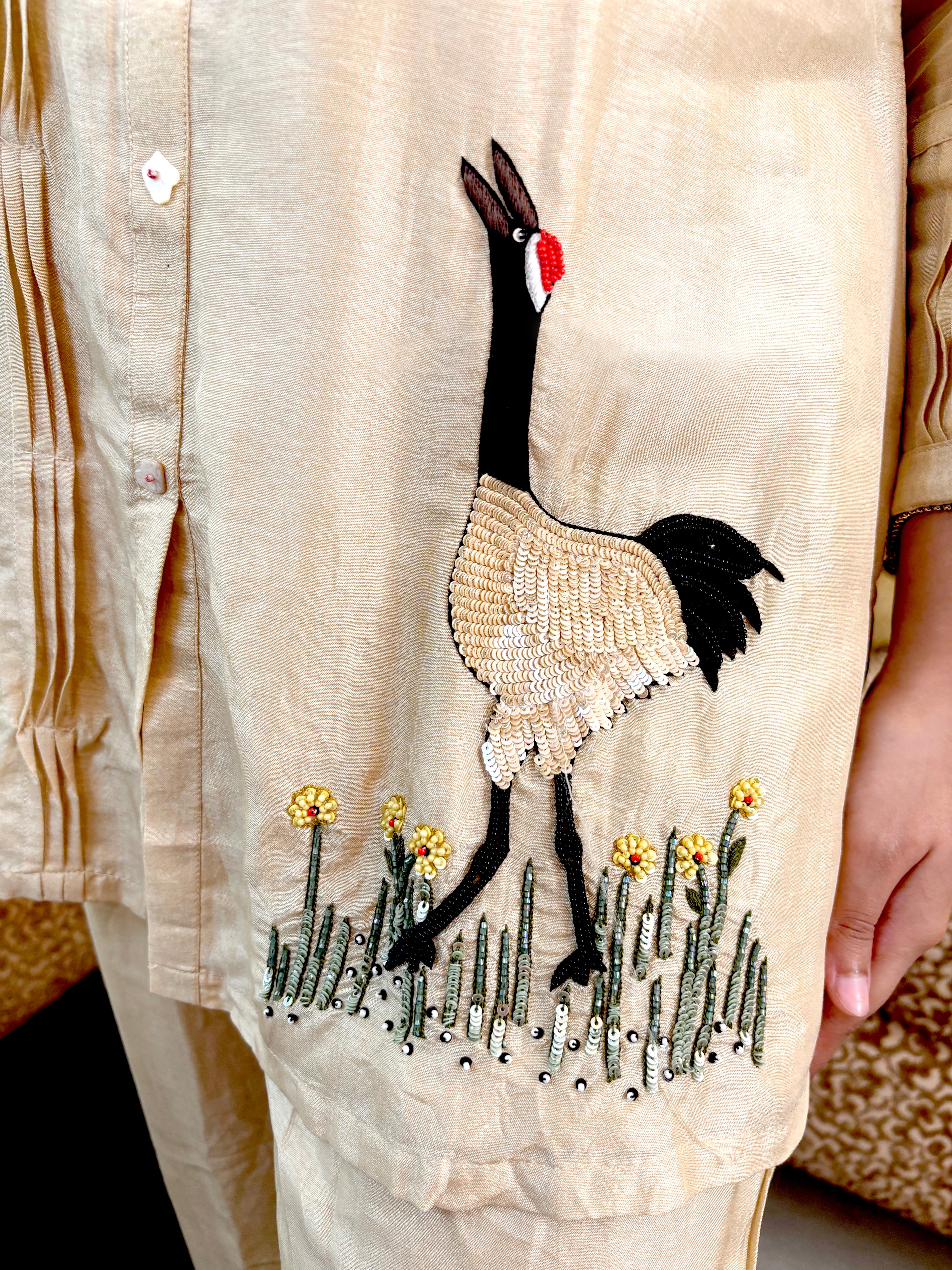 A close-up of a beige embroidered tunic or kurta featuring a large crane motif with black, beige, and red details, along with small yellow flowers and green grass at the hem. A person's hand is visible on the right side.