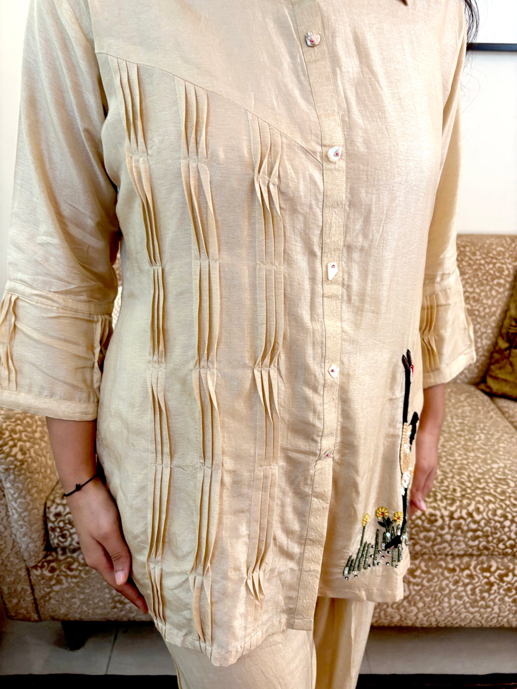 A person wearing a light beige or cream-colored kurta with vertical pleated detailing on the front and a small embroidered motif near the hem, standing in front of a patterned sofa.
