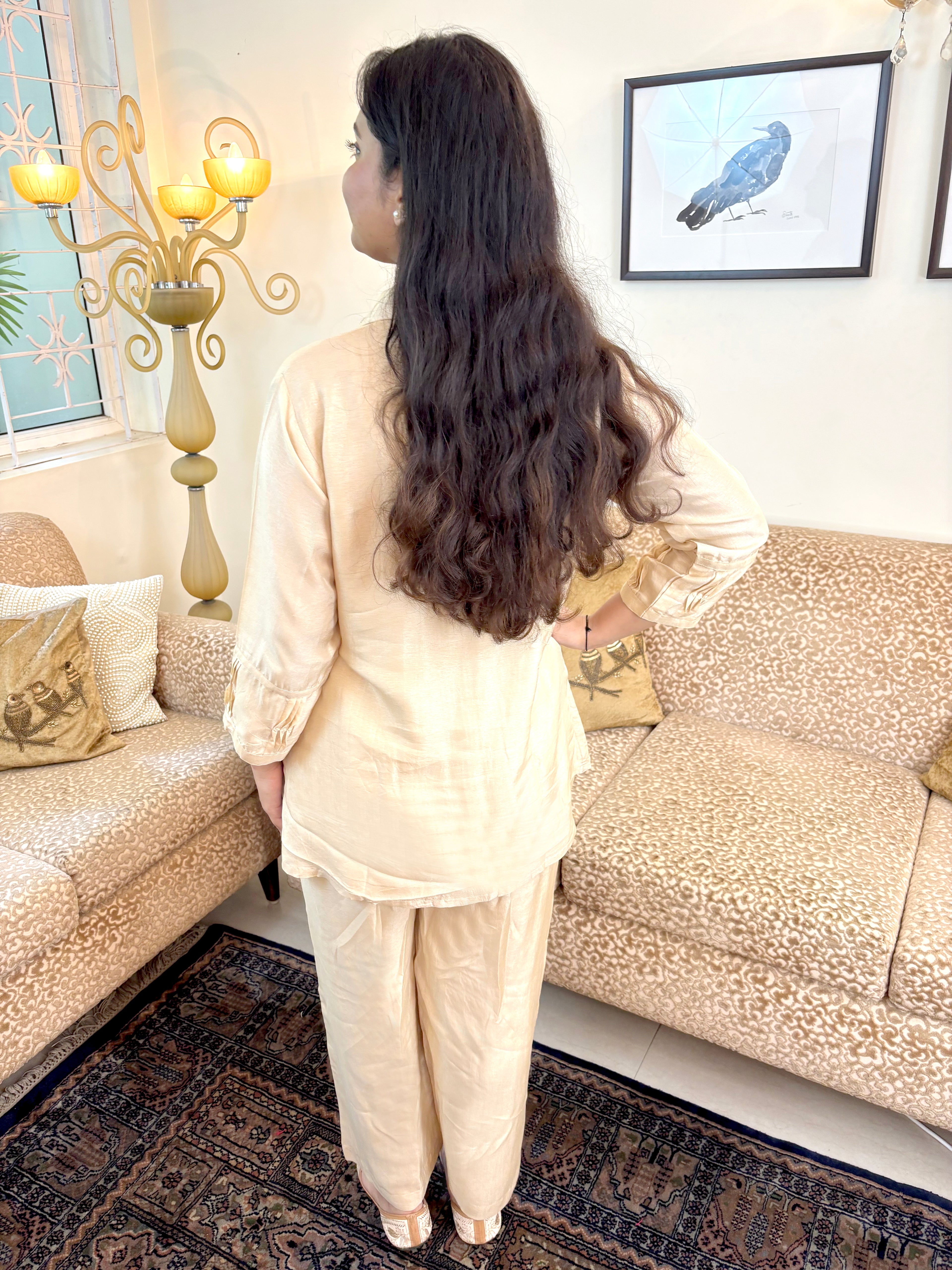 A woman with long, wavy dark brown hair is shown from behind, wearing a light beige two-piece linen pajama set in a living room setting with patterned sofas and a rug.