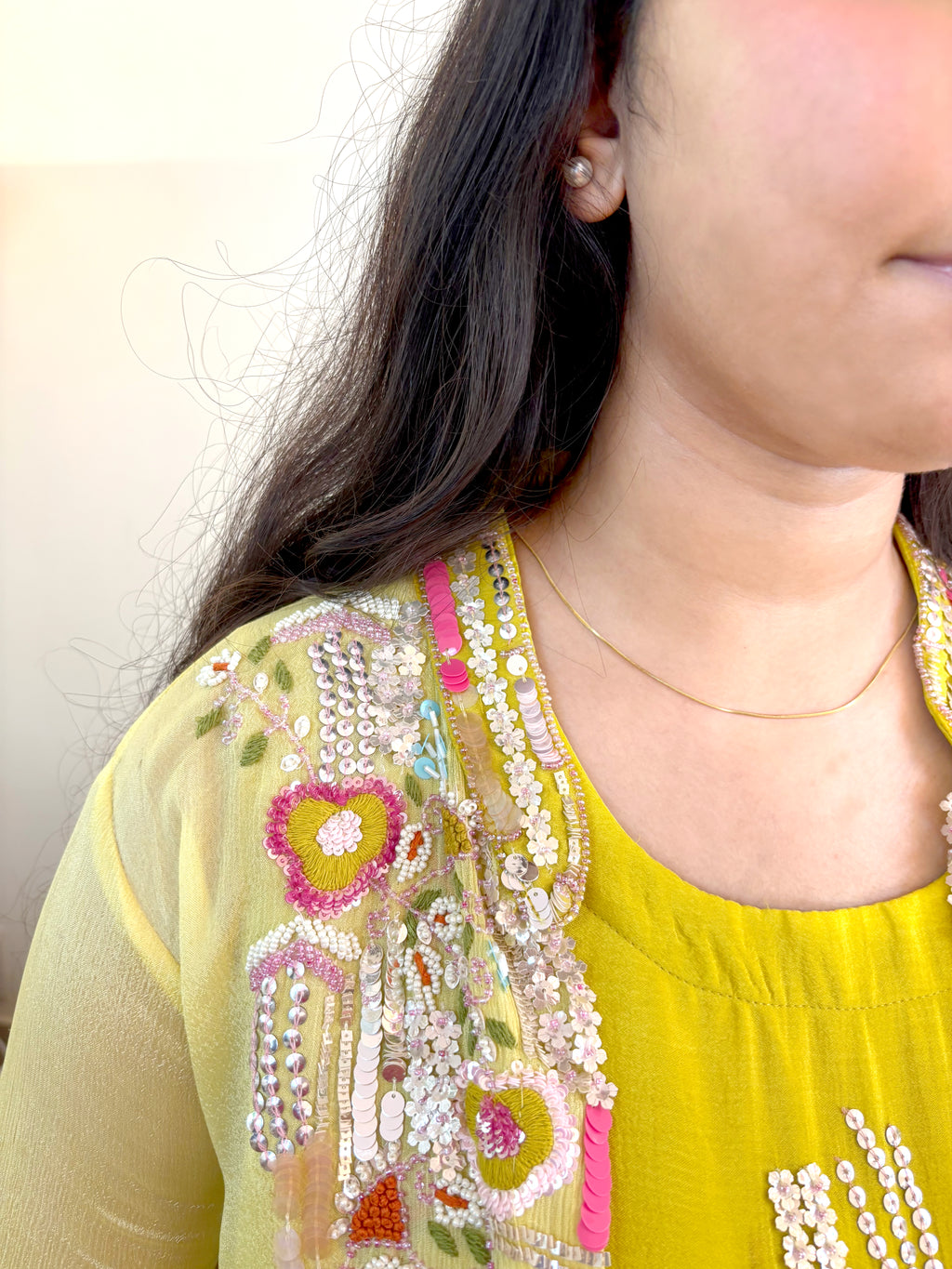 Close-up of a person wearing a bright yellow top with elaborate multicolor floral embroidery and beadwork on the neckline, along with a thin gold necklace.
