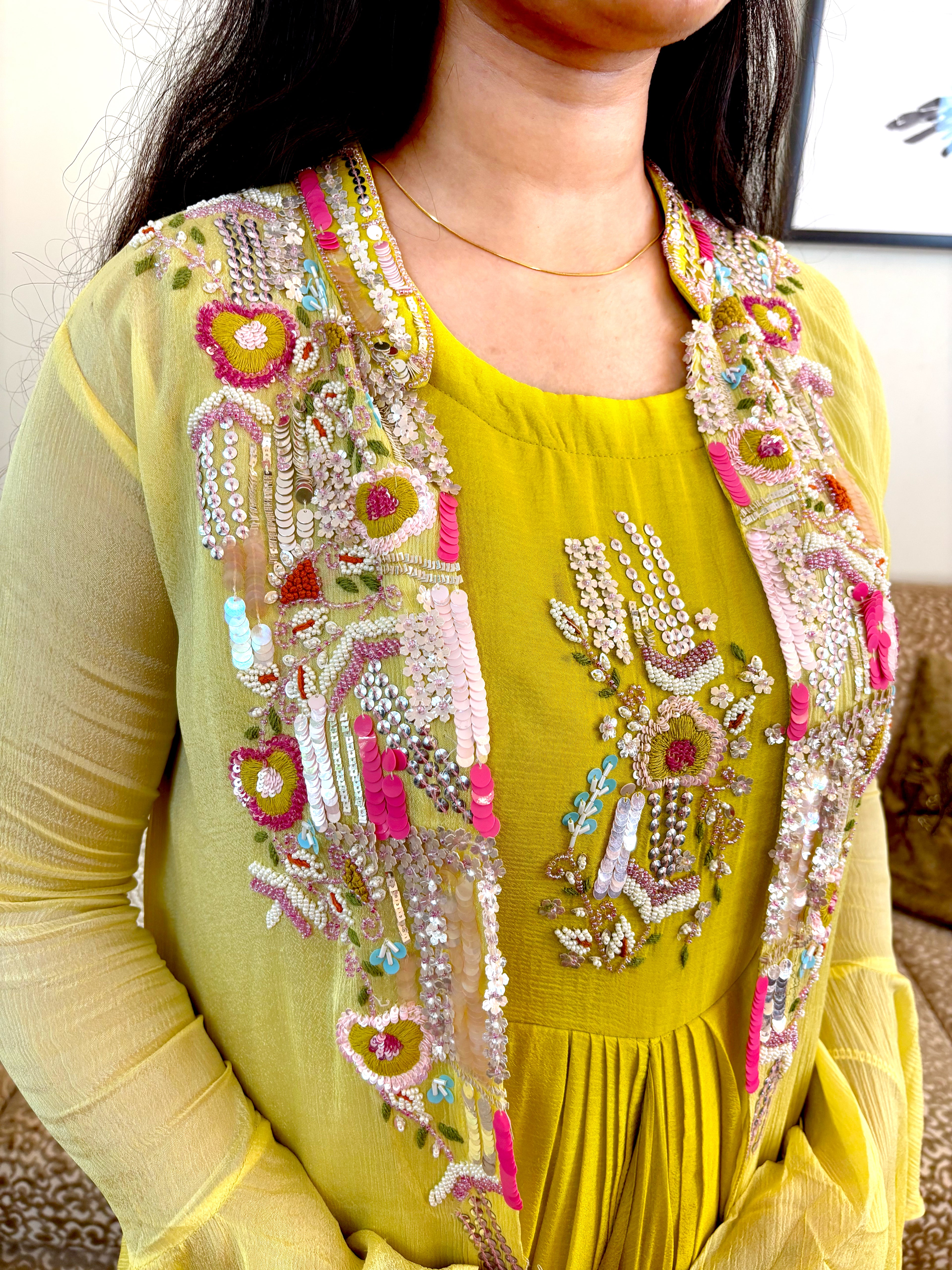 A close-up of a person wearing a bright yellow traditional outfit with an open, heavily embellished sheer vest featuring pink, green, and silver embroidery and sequins.